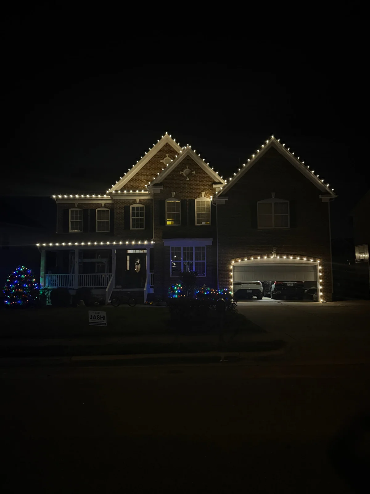 Warm white lights on the roof gables of a brick two-story home with multicolor lights on the front yard bushes and small trees